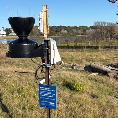 An RX2100 weather station mounted to a metal stake with a black rain bucket.  Mounted to the stake is a blue sign stating WATER QUALITY MONITORING STATION among other things.