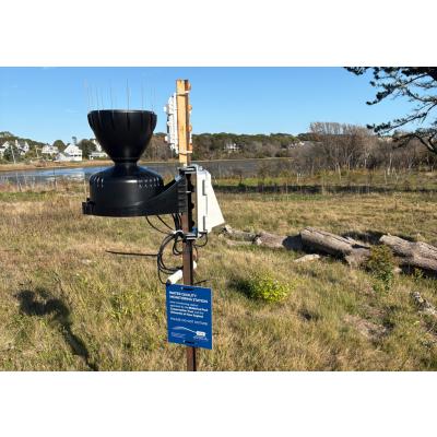An RX2100 weather station mounted to a metal stake with a black rain bucket.  Mounted to the stake is a blue sign stating WATER QUALITY MONITORING STATION among other things.