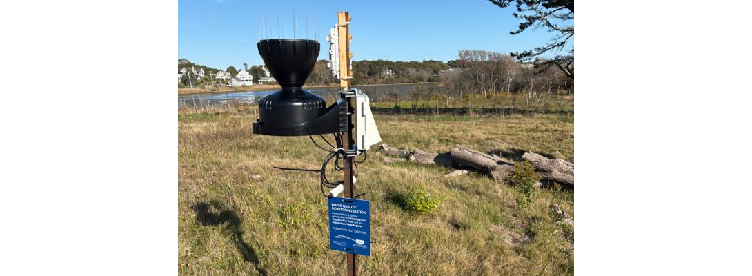 An RX2100 weather station mounted to a metal stake with a black rain bucket.  Mounted to the stake is a blue sign stating WATER QUALITY MONITORING STATION among other things.